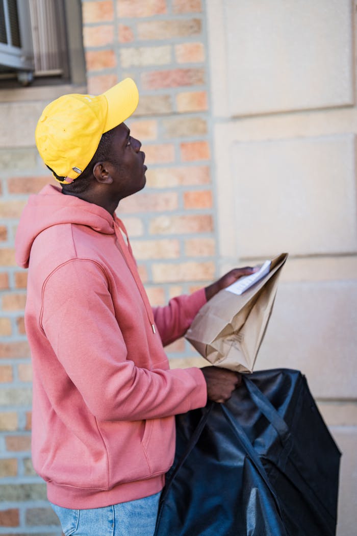 A delivery man wearing a yellow cap and hoodie holding a bag and parcel outdoors.