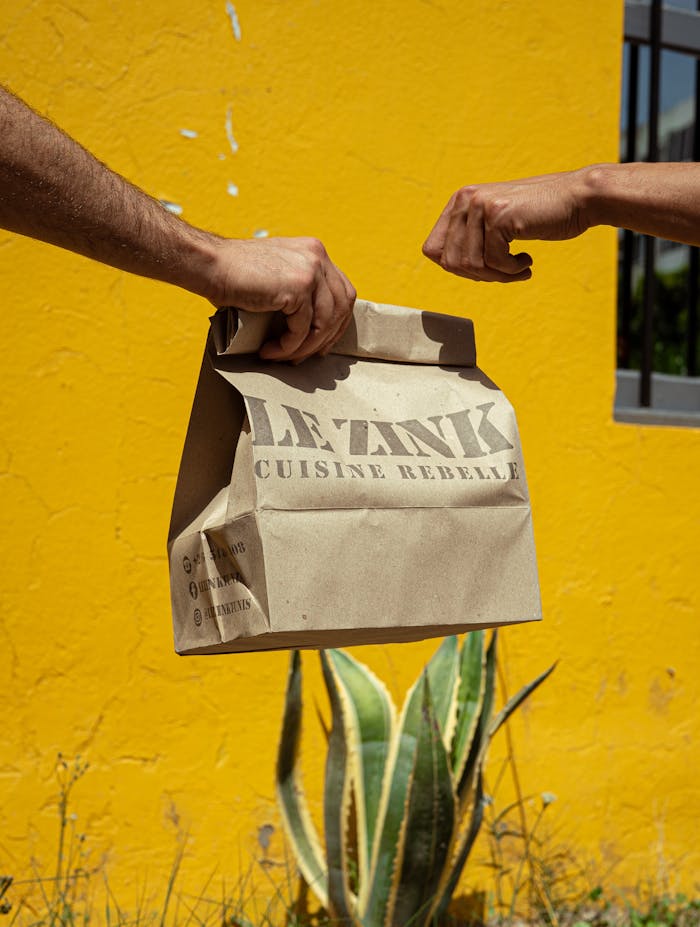 A paper bag hand-off in front of a bright yellow wall symbolizes sustainable urban food delivery.