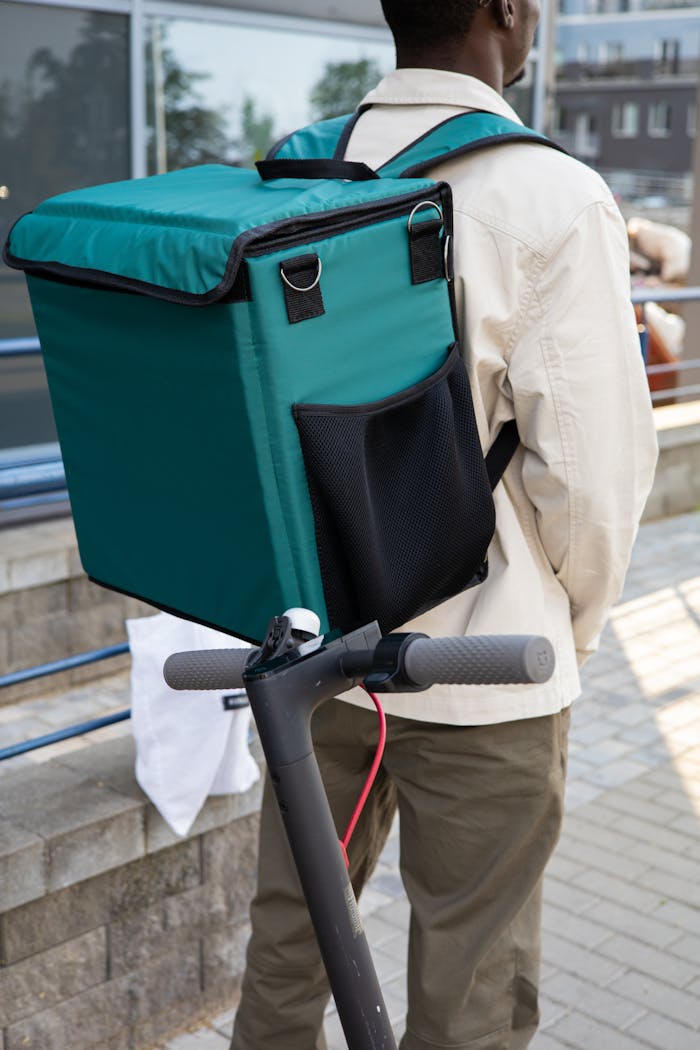 A delivery man riding an electric scooter with a thermal backpack outdoors.